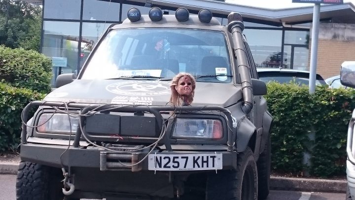 Badly modified cars thread Mk2 - Page 85 - General Gassing - PistonHeads - The image showcases a large grey military-type truck parked in a parking lot. The truck features a prominent grille and what appears to be heavy off-road capabilities. A woman is peeking out from behind the grille, adding a touch of human element to the scene. The license plate of the truck is clearly visible, reading "XKH." The truck is parked amidst other vehicles, suggesting the location might be a public parking area.