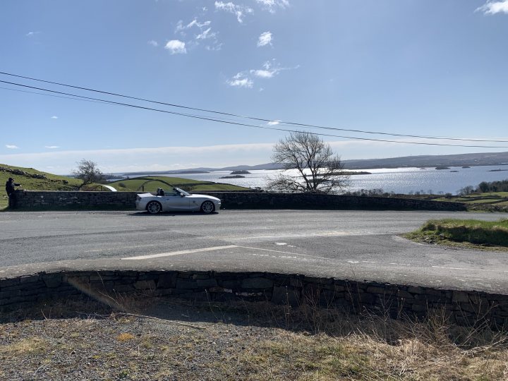 Southern Ireland 7 day driving tour. - Page 1 - Holidays & Travel - PistonHeads UK - The image captures a serene landscape dominated by a large, still lake. A car is seen driving on the right side of the road that stretches out into the distance. The sky above is clear and blue, suggesting a bright day. On the left side of the road, there's a stone wall which adds to the rustic charm of the scene. In the far distance, a small hill or land formation can be spotted on the other side of the lake.