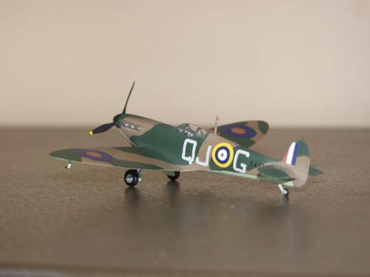 Spitfire September Pistonheads Scale - The image showcases a small model of a World War II fighter plane, specifically a QIitter eveneagle with a visible number "H" on its side. The plane is predominantly green, adorned with yellow and white markings, and three red and white circles. It's equipped with two propellers and sits on a flat grey surface. The model maintains its details, suggesting a sense of realism even at a small scale.