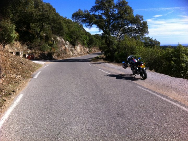 A man riding a motorcycle down a dirt road - Pistonheads - The image captures a dynamic scene of a single motorcyclist navigating a winding mountain road. The cyclist, clad in a black jacket and riding a black motorcycle, is leaning into a sharp turn, highlighting the action and motion of the ride. The road itself is nestled between the sky and a verdant landscape, with patches of grass and trees flanking its sides. The background reveals a breathtaking view of the sky merging with the vast expanse of a forested mountainside, adding a sense of adventure and freedom to the image.