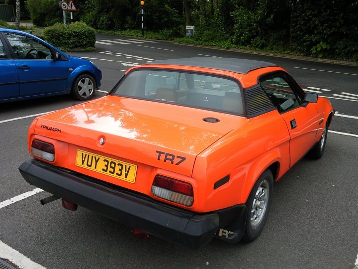 COOL CLASSIC CAR SPOTTERS POST!!! Vol 2 - Page 145 - Classic Cars and Yesterday's Heroes - PistonHeads - The image showcases an orange, vintage sports car parked in a parking lot. The car appears to be a Triumph TR7 sports model and is prominently positioned in the foreground. The surrounding environment consists of a blacktop road with traffic signs and a tree-lined street in the background. Other parked cars of different models and colors are also visible in the vicinity, adding to the urban setting.