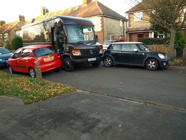 The BAD PARKING thread [vol3] - Page 463 - General Gassing - PistonHeads - The image depicts a humorous scene on a residential street. A large UPS truck occupies the right side of the frame, its presence seemingly commanding due to its size. On the left, other parked cars are dwarfed by this towering vehicle. Sandwiched between the towering UPS truck on the right and the compact Smart car on the left is a Mini Cooper, adding a stark contrast in size. The setting appears to be a sunny day, with the cars parked on a driveway. The image plays on the exaggeration of scale, creating a comical juxtaposition of the different vehicles.