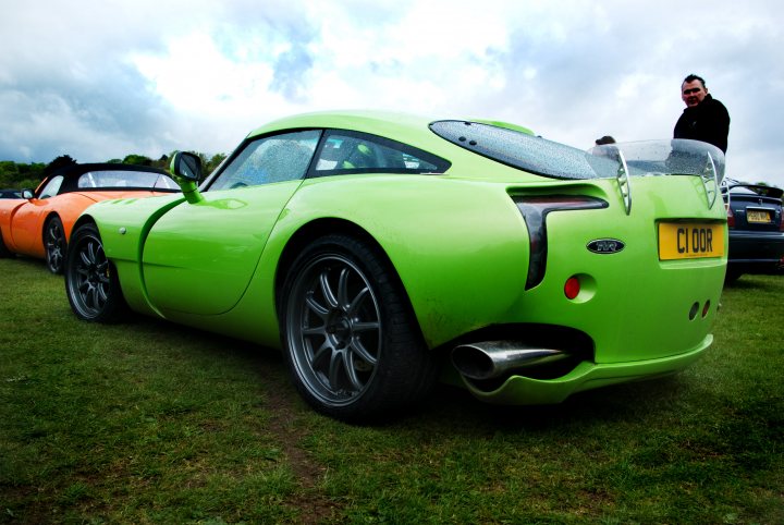 Pistonheads Fastlane - The image showcases a line of three sports cars parked in a grassy area against a backdrop of a cloudy sky. The car in the foreground is a vibrant lime green model with a distinctive sloping rear fascia. Among the reserve cars, there is a noticeable variation in color: a striking orange and a conservative black vehicle. All cars are neatly parked, looking ready for an event or excursion.