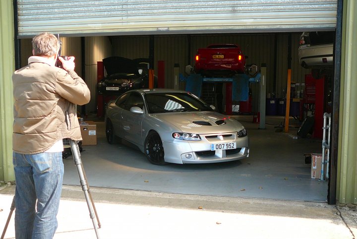 Pistonheads - In the image, a man is standing outside a garage, capturing the scene with his camera. The garage is open, revealing a parking lot where several cars are parked. Notably, there's a silver car parked in the garage and raises, suggesting some upcoming maintenance or repair. The man's focused posture indicates he might be a professional or a passionate car enthusiast, and he's aiming at the cars in the garage for a photo.