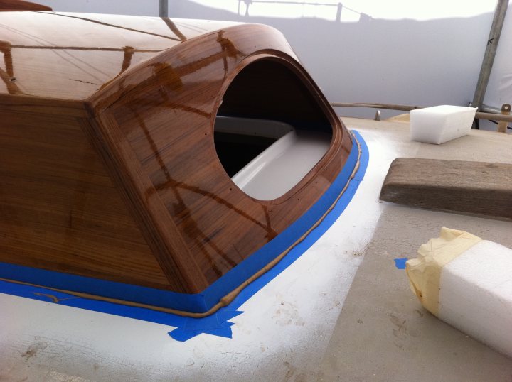 My 1968 Olsen Nimbus restoration project. - Page 18 - Boats, Planes & Trains - PistonHeads - The image shows a close-up view of a wooden boat being worked on in a workshop. The focus is on the bow, which is being repaired or constructed. The edges of the wooden panels are taped with blue tape, indicating an area of ongoing work. There's also a brick, possibly used as a support or trivet, and another piece of wood nearby. The background is slightly blurred, with hints of other structures, suggesting that this is a larger shipbuilding or construction environment.