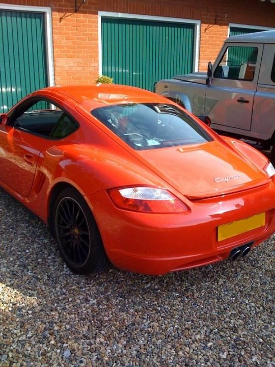 Pistonheads - The image shows a vibrant orange sports car parked on a gravel driveway. The car features a sleek design with a drop-top convertible roof and black rims that add to its sporty appearance. The vehicle is parked in front of a structure with green shutters, providing a contrasting background to the car's bright color. There is also another vehicle, possibly a truck, partially visible to the right.