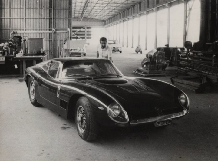 A 'period' classics pictures thread (Mk III) - Page 41 - Classic Cars and Yesterday's Heroes - PistonHeads UK - The image is a black and white photograph of a classic car. It's an old model car, possibly a muscle car given the design elements like long hood and large rear wing. The car appears to be in good condition, suggesting it might be well-maintained or restored.

The setting of the photo seems to be inside an industrial or warehouse environment, as indicated by the presence of metal structures and other machinery in the background. There is a person visible at a distance, possibly a worker given their attire, but they are not the main focus of the image.

There are no distinctive branding or identifying marks on the car that would allow for easy identification. The style of the photograph suggests it may have been taken during a time when color photography was less common or when the car's owner wanted to capture a timeless, classic aesthetic.