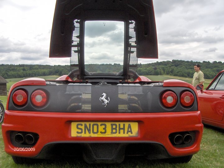 Rally Raleigh Pistonheads - The image captures a scene at what appears to be a classic car meetup. The main focus is a red, black, and yellow Ferrari F430 convertible with its roof up. The license plate reads "SN03 BHA". In the background, there's another classic car on display, and a tree-covered hillside can be seen under a partly cloudy sky. There's also a person in a beige shirt standing on the right side of the frame, adding a human element to this setting. A distinct date "20/05/2009" is displayed at the bottom left of the frame, suggesting the date when this photo was taken.