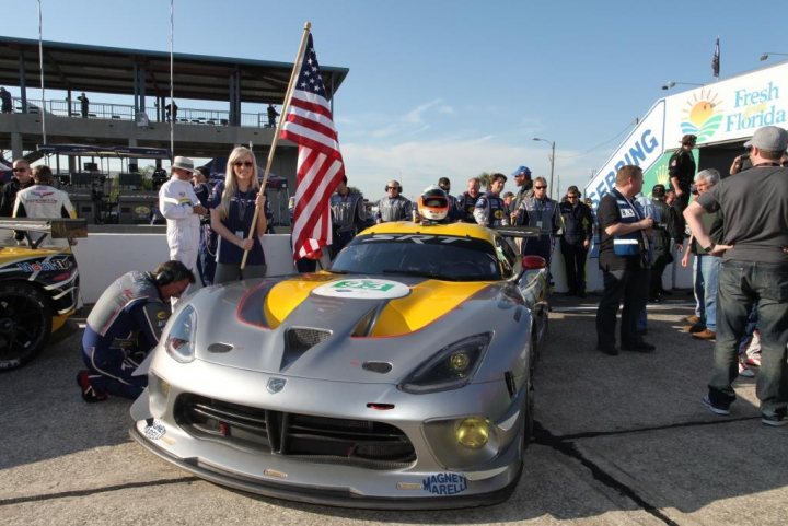 New SRT Advert - Page 1 - Vipers - PistonHeads - In the image, a vibrant racing scene unfolds in the sunlight. A crowd of spectators is gathered around several racing vehicles on display. The most striking feature is a race car with a yellow and grey color scheme, prominently displaying a large american flag at the front.