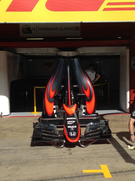 McLaren's new livery - Page 3 - Formula 1 - PistonHeads - The image features a Formula 1 car with a predominantly orange and black color scheme, prominently backed by sponsors. The car is parked in a covered garage within a track setting. A person is standing behind the front wheel of the car, wearing a dark shirt and shorts, possibly an attending mechanic or technician. There is an edition number "14" visible on the inside of the garage structure, indicating a specific location or being associated with a respected mechanic named Fernando Alonso. The area is brightly lit, suggesting it's possibly daytime due to reflections on the windows. The photo captures the realism of the sport with its technical precision and high-quality design.