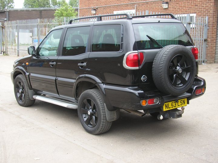 tuned terrano 3.0 di sve - Page 1 - Readers' Cars - PistonHeads - The image displays a black SUV parked on a tarmac area. The vehicle is a modern design with a spare tire mounted on the back door. It features raised suspension and five-spoke alloy wheels. The background indicates a commercial or industrial setting, with a chain link fence and a brick building visible. There is no visible text or branding on the vehicle or in the image. The photo appears to be a stock image rather than a specific listed car.