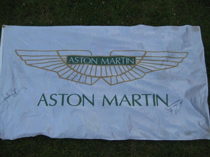 Merci les Belges/KRONOS - Page 1 - Aston Martin - PistonHeads - The image features a large flag with the emblem of 'Aston Martin' prominently displayed in the center. The emblem is golden and includes a bird design. The flag has additional gold text that reads 'Aston Martin'. There are signatures on the flag, presumably of people associated with the brand. The flag is laid out on a grassy surface. The text and branding suggest that the flag is likely a piece of merchandise or a decorative item.