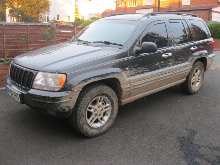 Pics of your offroaders... - Page 41 - Off Road - PistonHeads - The image shows a dark-colored SUV parked on an asphalt surface. The front of the vehicle is angled toward the viewer and the photo is taken during the daytime. The SUV has visible dirt, mud, and dust on its exterior, suggesting it has been on rough terrain or has accumulated dirt over time. There is no additional context or surrounding environment provided, such as houses or the sky. The SUV has alloy wheels and there appears to be a mirror visible on the driver's side.