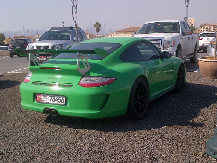 Middle East spotted thread - Page 64 - Middle East - PistonHeads - In the image, a vibrant green Porsche is parked on a gravel lot. The Porsche is positioned in such a way that its rear end is facing the viewer. The car is sports car model with a prominent rear spoiler. In the background, there are tall buildings and more cars parked, suggesting this could be in an urban area. The sky appears overcast, casting a soft light over the scene. The Porsche is parked next to a lamp post, adding a touch of street lighting to the outdoor setting.