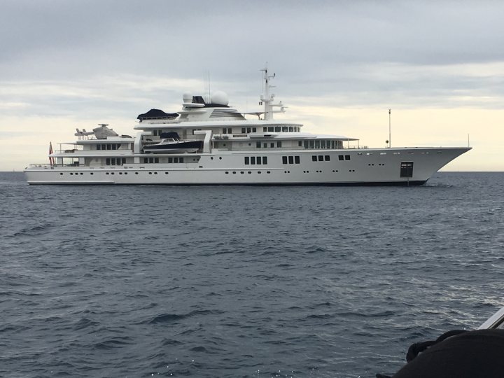 super yachts 60million+ - Page 189 - Boats, Planes & Trains - PistonHeads - The image shows a large, white luxury yacht with multiple decks, floating on calm water under an overcast sky. The yacht has numerous windows and is equipped with what seems to be a helicopter pad on the roof. A small skiff is visible in the foreground, with its bow facing towards the yacht. The boat is moored with a line tied to a cleat, indicating it's not moving.