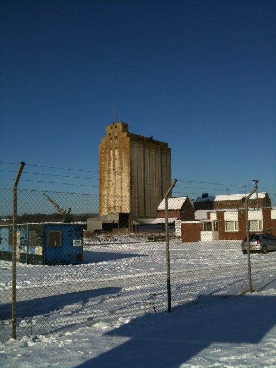 Ruins Pistonheads Nsomerset - The image captures a desolate industrial area, where time seems to have left its mark. A tall, brown grain silo stands as a monument to a different era. Its flat roof contrasts with the white, snow-covered top. To the right of the silo, a blue storage container is frozen in place by the chill of the winter's day.

An iron fence stretches across the foreground of the image, partially obscuring our view of the silo. Beyond the fence, houses can be seen blanketed in snow, their muted colors adding to the sense of stillness.

Despite the cold, the scene is not completely devoid of life. A single car, parked haphazardly, bears testament to the presence of human activity. Amidst the monochrome palette, this car introduces a splash of detail and adds a touch of life to this wintry tableau.