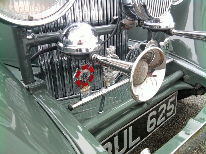 Saw this today... - Page 1 - Classic Cars and Yesterday's Heroes - PistonHeads - This image captures a close-up view of the grill and chrome fender of a vintage vehicle. The grill features a round emblem with the letter "B" and additional text, possibly indicating a make or model. The chrome fender on the side is adorned with a large red and white star. The vehicle's color is not prominently visible, but it's green with white letters. The shine of the chrome emphasizes its vintage style. The image is taken from a low angle, providing a detailed view of the vehicle's front.