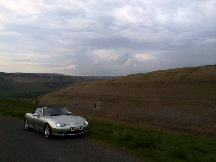 my mx5 - Page 1 - Mazda MX5/Eunos/Miata - PistonHeads - The image depicts a serene landscape featuring a grey convertible parked on the side of a narrow paved road. The road gently ascends, offering a clear view of a large, grassy hill in the distance. The upper layers of the cloudy sky are a blend of grey and white, creating a subdued backdrop for the scene. The convertible, with its sleek design, adds a touch of modernity to this tranquil rural setting.