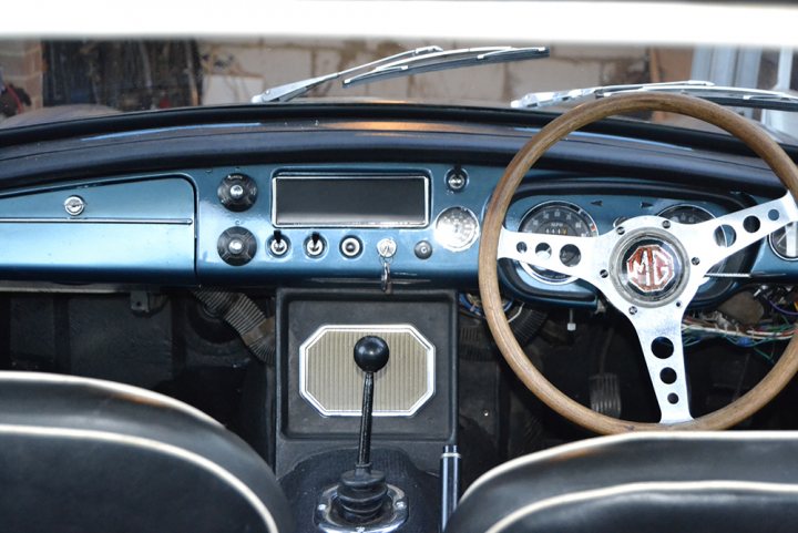 1966 MGB GT - stolen - please help! - Page 1 - MG - PistonHeads - The image showcases the interior of a vintage car, focusing on the steering wheel and dashboard area. The steering wheel, which is centrally located, is wooden with pebble-like patterns around the center ridge. The dashboard exhibits a blue and black color scheme with silver or chrome accents, and there's a mix of circular and rectangular shapes that characterize the vintage aesthetic. The steering column curves gently, and there's a gear or throttle pedal visible. The image provides a sense of nostalgia and history, particularly if the viewer has an interest in classic or vintage vehicles.