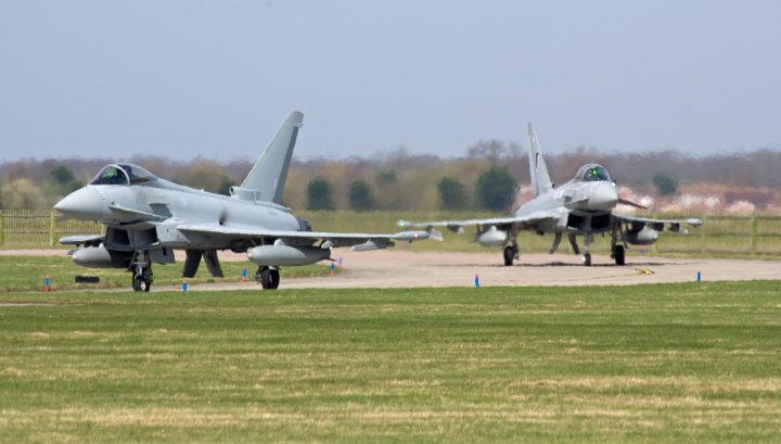 Cosford Pistonheads Airshow Aircraft - The image depicts an airfield with a clear sky. There are two fighter jets, one towards the top left and the other towards the middle right, arranged on the ground with visible landing gear. Surrounding the jets are a few people, and there are portable stairs parked next to the aircraft, indicating that they are potentially in the process of boarding or disembarking. A few trees can be seen in the background, beyond the runway area.