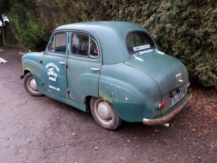 The Humer Unbeam Interesting Filling Stations Thread - Page 36 - General Gassing - PistonHeads - The image features an old, green vintage car parked on a gravel driveway. The car has a noticeable rust and dirt on its body, giving it a weathered appearance. It's missing the right side headlight and side rear window, and the trunk lid is also dented. The vehicle has a sign on the back that reads "sporting car service" and another one underneath that says "Jay's custom car". The background is overgrown with shrubs and greenery, suggesting that the car has been stationary for quite some time. The setting appears to be in a rural or semi-rural area, away from heavy traffic and metropolitan environments.