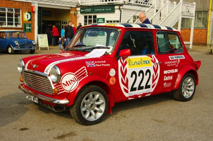 Mini Brooklands Day Pistonheads - The image features a striking red Mini Cooper, its front dominating the frame. The car is adorned with numerous decals and signs. On the hood, the words "LINCOLN MOTORS" and "ANDY BUSHEL" are prominently displayed, suggesting the car might be part of a dealership or used for promotional purposes. There are also mentions of "MICHEL VON HEINTZEN", which might refer to the car's owner or company involved in its use. Furthermore, a number "221" is visible, which could indicate a registration or racing number. The car is parked in a lot with a building and a sign that reads "PARKING ONE" in the background.