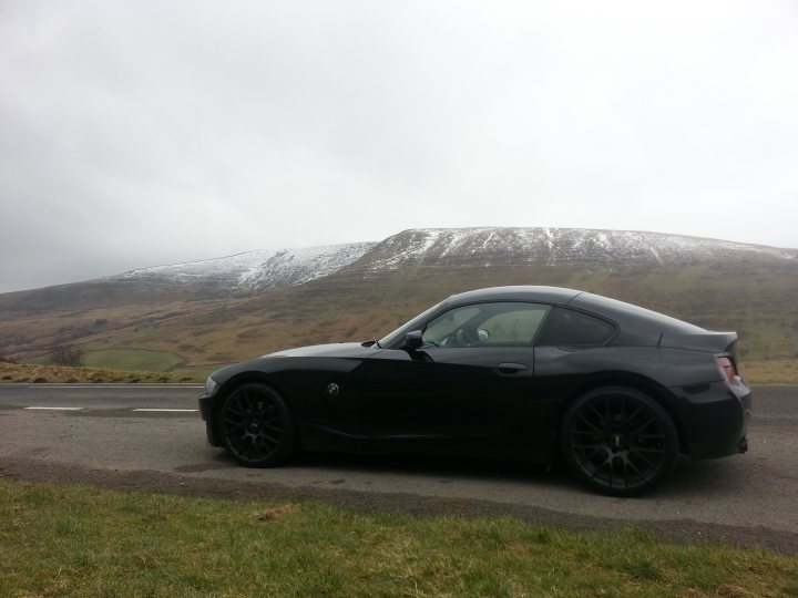 Decent South Wales drives - Page 1 - South Wales - PistonHeads - The image features a black sports car parked on the side of a country road, bathed in the early morning sunlight that casts soft shadows and highlights the car's sleek design. The car is positioned in profile view, pointing towards the left side of the photo where the road curves gently. The background is a serene pastoral landscape of rolling hills, sparse trees, and a distant mountain range covered in snow, providing a stark contrast to the aesthetically modern vehicle.