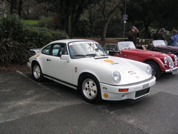 A car is parked on the side of the road - Pistonheads - In the image, an ancient white Porsche with a prominent badge is parked in a lot. The car sports a black soft top and has a round emblem on the bonnet. The lot is adjacent to a forested area, suggesting a rural or semi-urban setting. In the background, some people can be seen, adding to the everyday atmosphere of the scene. A red car is also visible in the background, providing a vivid contrast to the white Porsche.