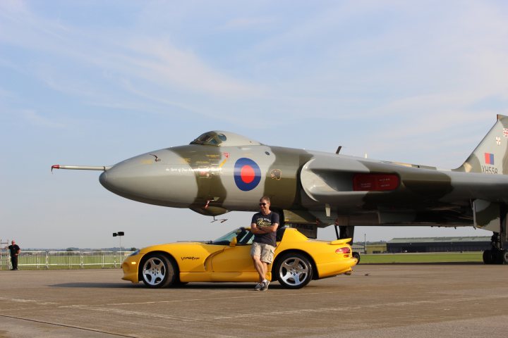 Pistonheads - The image depicts a striking scene on an airfield. The main subject is a camouflaged fighter jet, its wings spread wide, parked in a prestige position on the tarmac. A swaggering individual, clad in sunglasses, is casually leaning against the nose of the yellow sports car parked directly in front of the jet. In the background, another fighter jet is on display, adding depth to the scene. The third fighter jet is parked further back, providing context to the setting. The overall atmosphere is one of confidence and luxury.