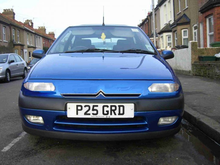 The image shows a blue car parked on the side of a road. The vehicle has a license plate that reads "P25 GRD". It appears to be a two-doored car. In the distance, there are residential properties, indicating the location is likely a suburban area. The sky is overcast, suggesting it might be a cloudy day. The street is relatively quiet with no visible traffic.