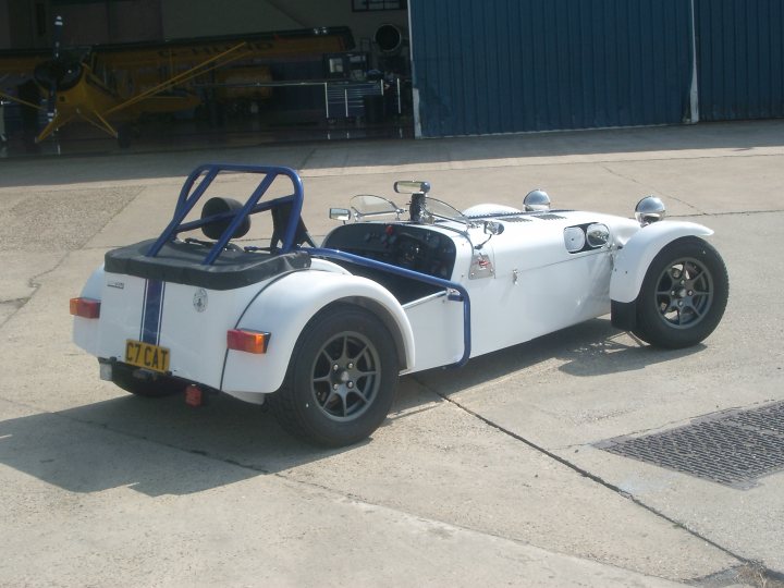 Pistonheads - This is an image of an old-fashioned, single-seater vehicle, not a car but a small tractor. It appears to be a 'Cat' brand vehicle, as indicated by the yellow license plate. The tractor is white with black tires and wheels, and the seat is blue. It's parked on a tarmac next to a building with a blue door. The tractor is equipped with a steering wheel at the front and a raised platform with a railing behind the seat for the driver's convenience. The vehicle's overall condition suggests it's well-maintained and ready for use. The small tractor seems to be the central focus of this image.