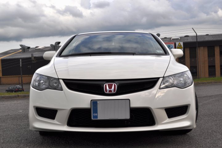 Import Type Pistonheads - The image shows a white Honda car with its headlights on, parked on the side of a road. It appears to be an overcast day, as indicated by the cloudy sky in the background. The car is equipped with a rear spoiler and is ready to merge into traffic. In the distance, partially obscured behind the vehicle, there's a yellow traffic sign.