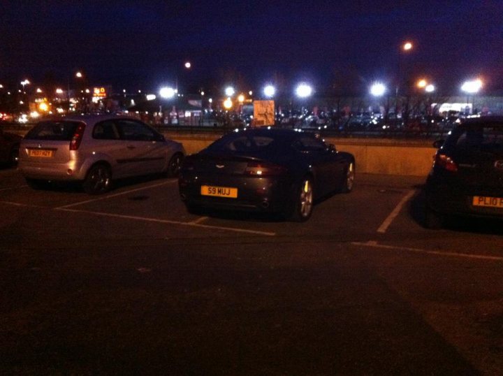 Pistonheads - The image captures a typical urban night scene, featuring a large, concrete parking lot bathed in artificial light. Two cars, a small silver hatchback on the left and a black sports car on the right, are parked side by side, their details subtly illuminated. The parking lot is situated against a backdrop of commercial buildings, their facades dark and unadorned under the gloom of the evening sky. The asphalt's smooth texture is evident under the artificial glow, reinforcing the stillness of the scene. Despite the numerous visible parking spaces, the lot appears to be mostly empty. The overall atmosphere of the image conveys a sense of quiet tranquility.