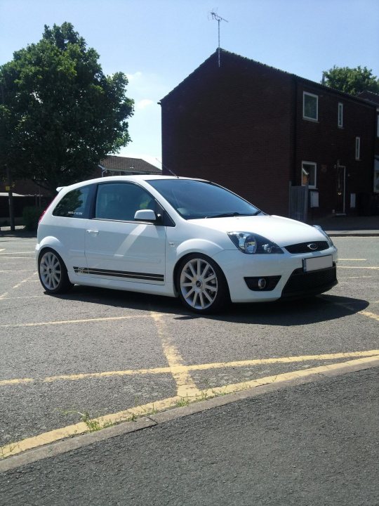 Alloy Pistonheads Refurbs - The image shows a white car parked at an angle on a street. The vehicle has five doors and a distinctive chrome strip running along its side. Behind the car is a two-story building with brick walls, and on the left side of the image, there is a tree with green foliage. The sky is clear and the lighting suggests it is a bright, sunny day. There is yellow marking on the road where the car is parked, which indicates a parking space. The car's wheels feature a design with multiple spokes.