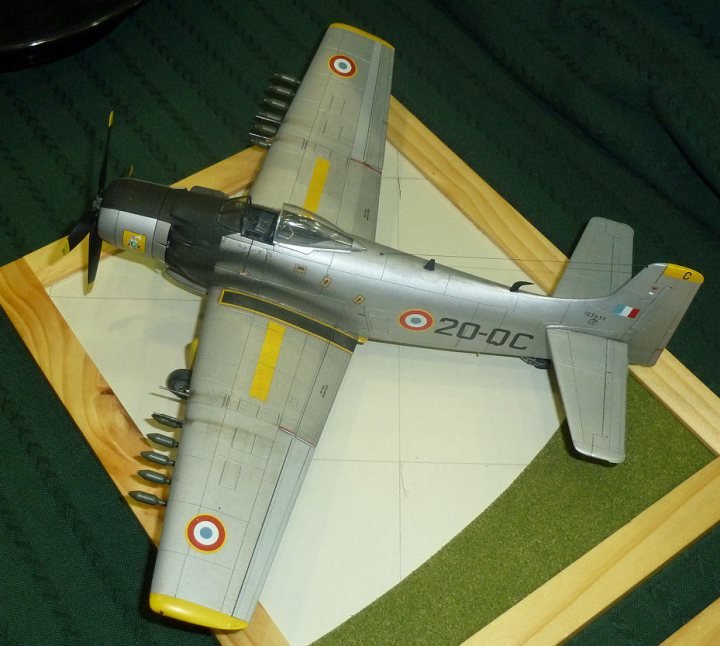 Pistonheads Model Crewe Feb - The image shows a detailed model of an airplane, specifically a fighter jet, captured in a moment of static display. The airplane, painted to resemble a military craft, is predominantly silver, with accents of yellow and orange. The model is set against a textured green background, which could provide a crude representation of a runway or a grassy field, further enhancing the realism of the display. The wooden frame holding the model itself adds an element of craftsmanship, reminding the viewer of the skill involved in assembling and detailing a model like this. The name "AMRIT 20-DC" is visible, suggesting that this model might be associated with a specific event or group, possibly indicating participation in an air show or event.