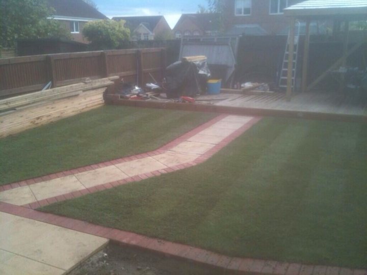 ideas for lawn /gravel edging ? - Page 1 - Homes, Gardens and DIY - PistonHeads - The image is a photograph of a lawn in the midst of being landscaped. The foreground shows a section of freshly laid artificial turf with a distinctive wavy pattern and red edging. Behind the turf, there are several tools and materials scattered on the ground, indicating ongoing work. There is a garden bench to the left side of the image, turned over as if accidentally knocked over in the midst of the job. The background features an outdoor structure, possibly an arrangement of wooden pallets, and there's a gray recycling bin. There are no people visible in the photograph.