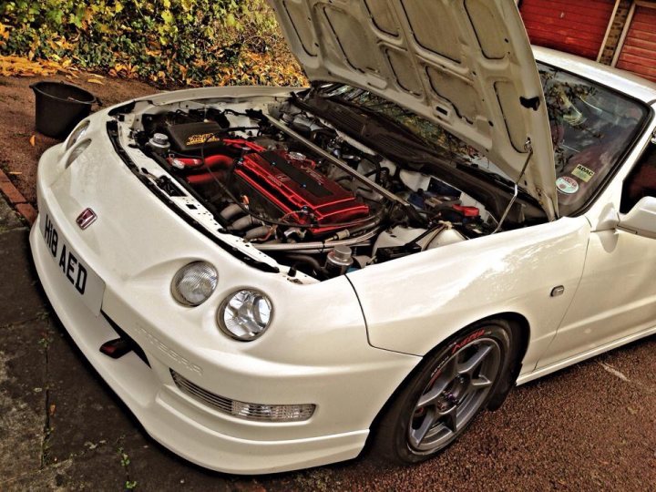 A car parked in a field with a lot of luggage - Pistonheads - The image shows a white car with its hood propped up on a driveway. The engine seen under the hood is quite intricate, featuring a colorful array of tubes and mechanical components, suggestive of modifications or a high-performance build. The front bumper of the car is intact and there's a noticeable curvature to the car's body, indicating a possible aftermarket styling. The surrounding environment hints at a residential area, possibly the car's home driveway or a similar setting.