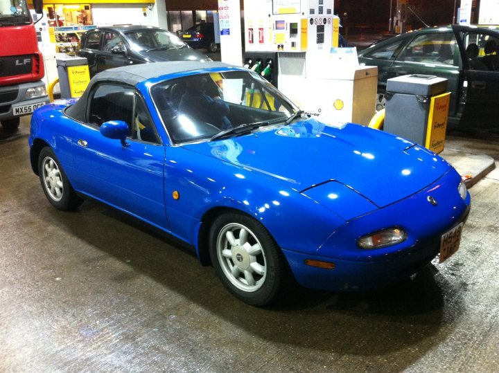 Yet Another MX-5! - Page 1 - Readers' Cars - PistonHeads - The image shows a vibrant blue sports car positioned in a gas station station. Its sleek design and shiny surface reflect the surrounding lights. The gas station is quite busy, with visible pumps and fuel price signs in the background. Other cars are also parked in the lot, indicating a public nature of the location. The overall lighting and ambiance suggest it might be evening or during the night as the environment is well illuminated.