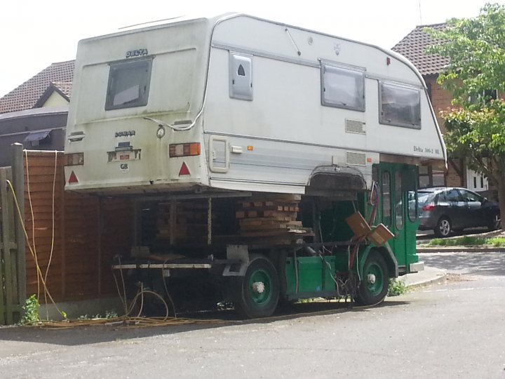 OMG...how stunning!.... - Page 1 - Tents, Caravans & Motorhomes - PistonHeads - The image shows a large truck with an attached trailer. The trailer is painted white and has a green rear section. On the side of the trailer, there are numerous crates and a pallet, with additional ground clearance provided by a large flatbed carriage. In the background, there are residential houses, and a driveway leads up to this vehicle, with various cars parked on it. The sky is overcast, suggesting an early morning or overcast day.