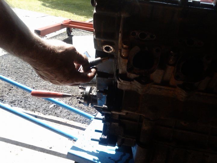 Engine Situ Timing Pistonheads Procedure Remove Cover Proposed - The image shows a close-up of a hand holding a screwdriver, engaged in a repair or construction activity. The screwdriver appears to be adjusting or removing a metal piece from a larger assembly. The screwdriver is angled towards the bottom left corner of the image, resting on a blue leverage bar. The background is blurred, but it appears to be an outdoor setting with a gravel ground, which might indicate a work site or garage. The focus of the image is on the hands and tools, suggesting a theme related to mechanics or DIY projects.