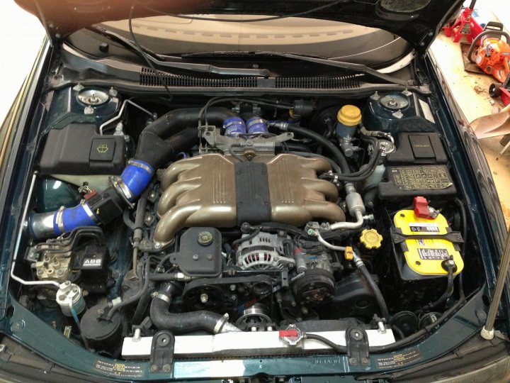 A close up of a luggage bag on a car - Pistonheads - The image shows an interior view of a small car, specifically the engine compartment. Various components are visible, including what appears to be the radiator, a blue pipe, wiring, pumps, and belts. Electrical components and hoses are installed next to the batteries. A yellow accessory case is also visible in the open engine compartment.