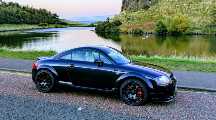 What do you guys think about the audi tt mk1 225? - Page 2 - Car Buying - PistonHeads - The image depicts a black Beetle car parked on the side of a road. The car has a sporty appearance with red brake calipers. The road is near a serene body of water, which reflects some trees and bushes nearby. The car is positioned with the front facing towards the water, and there's a clear sky above indicating either early morning or late evening, with the sun possibly on the horizon.