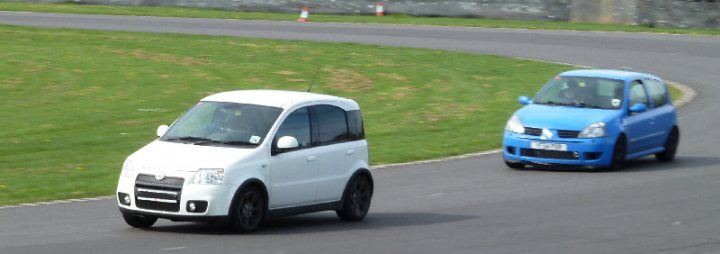 Pistonheads Combe Castle Tomorrow - The image shows two small cars driving on a road adjacent to a field. The car in the foreground is white with black wheels and is positioned closer to the camera, while the second car is blue and is seen as the car is moving away from the camera. The vehicles seem to be compact in size, potentially from a specific automotive brand, and the overall scene conveys a sense of motion. The setting appears to be an outdoor environment with open space. There are no visible passengers in the cars, suggesting the focus of the image is on the cars themselves and their movement.