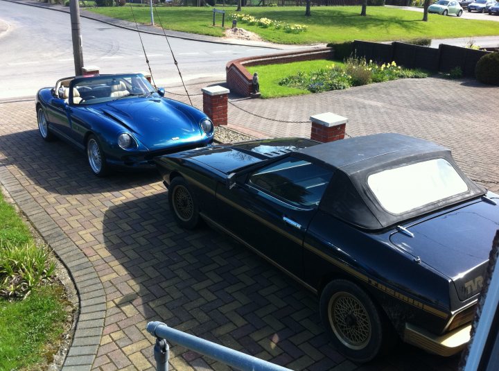 A car is parked next to a parking meter - Pistonheads - This is a colorful image of a well-kept residential driveway with two classic cars parked on a brick pavement. The car on the left is a blue convertible with the top up, featuring a wire-framed hood, which is adorned with various brand logos, including one that looks like a capital "S." The brick paving around the backyard has a distinctive pattern, and there's a wooden fence in the back. There's also a street visible with parked cars along the road. The atmosphere appears to be calm and residential.