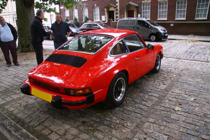 Queen Sq Tomorrow - Page 1 - South West - PistonHeads - In the heart of a cobblestone street, a bright red Porsche 911 GT3 RS, with its distinctive aerodynamic body, is parked. Two men in casual attire stand in front of it, one slightly closer than the other, appearing to be inspecting or admiring the vehicle. Behind them, further down the street, additional people and cars are visible, adding to the urban ambiance. The vintage appeal of the Porsche adds a touch of luxury and nostalgia to the scene.