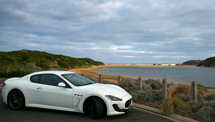 Great Ocean Road - Page 1 - Australia - PistonHeads - The image captures a serene scene by the seaside. A white Maserati coupe, sleek and stylish, is parked on a paved area adjacent to a fence. The car is facing the ocean, which stretches out in the horizon, meeting the sky at a distance. The foreground is dominated by a rocky landscape with vegetation adding a touch of greenery to the scene. The atmosphere is peaceful, with the sky appearing slightly cloudy, casting a soft light over the landscape.