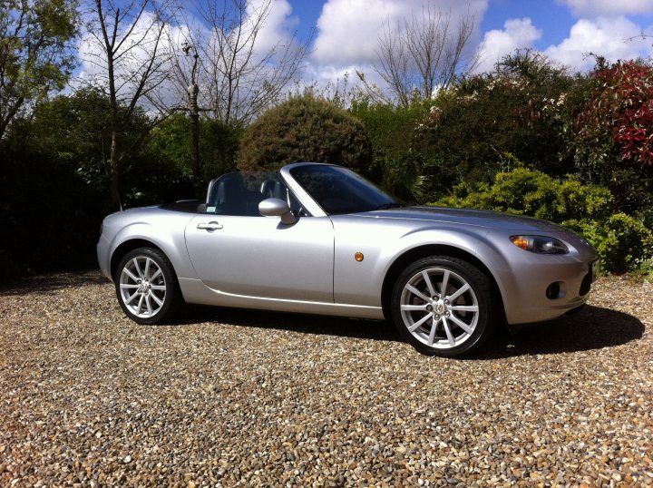 New and happy owner! - Page 1 - Mazda MX5/Eunos/Miata - PistonHeads - The image shows a silver two-door convertible sports car parked on a gravel surface surrounded by shrubbery and trees. The car features alloy wheels and is structured with a soft-top roof that's down. The sky overhead is cloudy, suggesting a potentially overcast day. The overall setting implies a suburban or rural environment, possibly in the early morning or late afternoon, given the soft lighting. The car appears to be well-maintained, further showcasing its sleek design and shiny exterior.