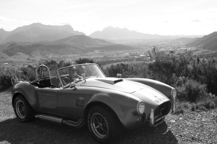 One of the most polite places I have driven - South Africahe - Page 1 - Advanced Driving - PistonHeads - The image is a black and white photograph capturing a historic race car parked in a hilly, expansive open area with a city landscape in the background and mountains beyond. The car is an old sports or racer, possibly Ferrari or a similar European marque. The driver's side door is open, adding a narrative element to the scene. The perspective of the photo suggests it was taken from a lower angle, looking upward towards the car, emphasizing its stature and presence. The landscape spans vast distances, reinforcing the open and expansive feel of the image.