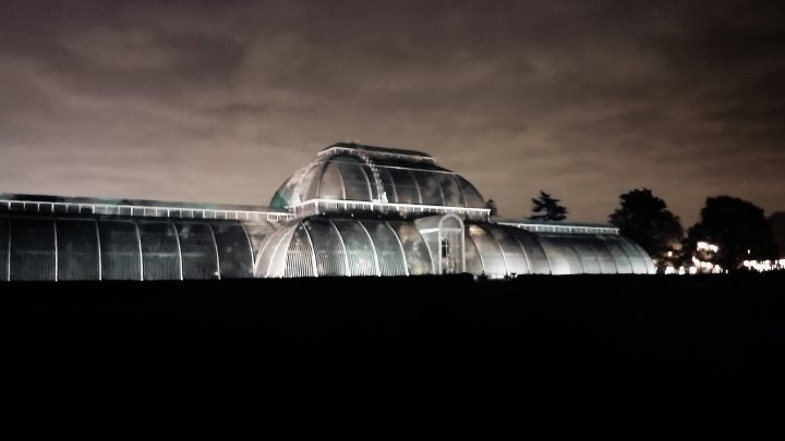 Just for fun random camera phone pictures you're happy with. - Page 9 - Photography & Video - PistonHeads - The image depicts a scene at twilight, with a large, metallic greenhouse or shed structure prominently featured against a dusky sky. The building has an arched shape with a curving roof, illuminated by internal lights. Visible around the perimeter of the illuminated structure are trees, indicating it might be located in a rural or semi-rural setting. The photograph has a color palette that emphasizes greens, blacks, and soft grays, conveying a sense of tranquility and evening serenity.