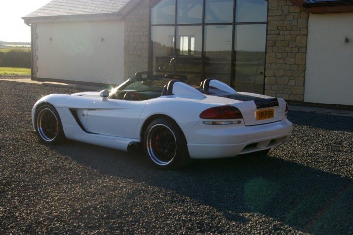 Blown But Not Blown - Page 1 - Vipers - PistonHeads - This image features a white sports car parked in front of a house. The car is positioned on the left side of the image, adjacent to a gravel driveway. Its top is down, suggesting a sunny day. The car appears to have a high performance model, indicated by the rear spoiler, which is prominently visible above the taillights. The setting sun casts a warm glow on the parked vehicle and its surroundings. The house in the background has a traditional architectural style, with a visible chimney and what appears to be a stone wall.